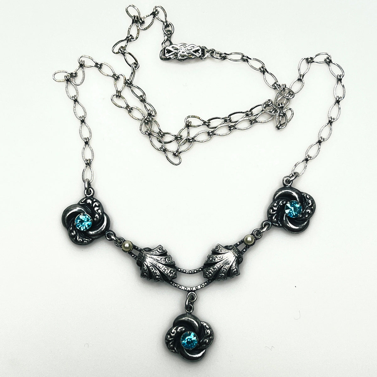 Close up of vintage silver-tone necklace showing dimensional floral ornaments, textured chain and central pendant.