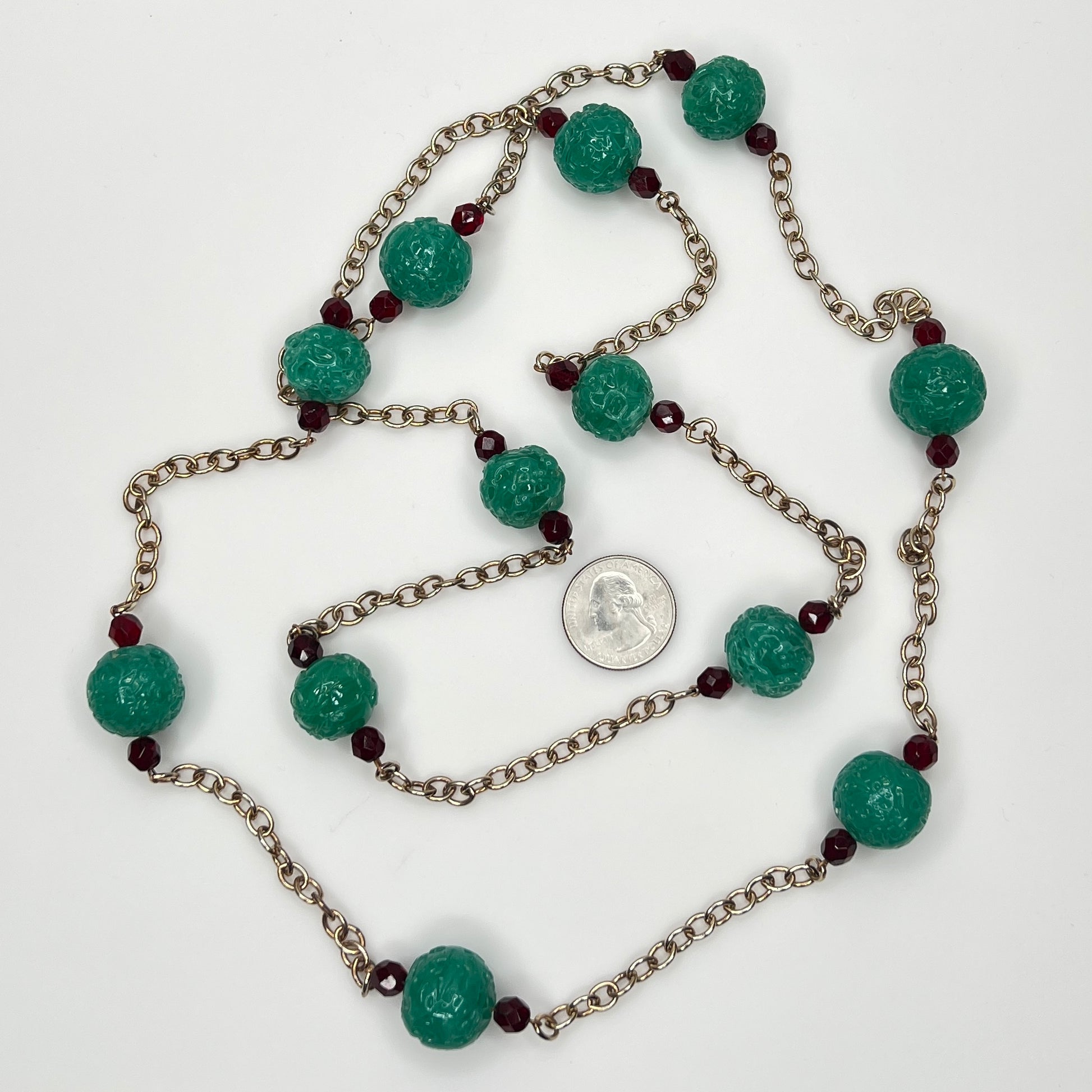 Close-up of textured green art glass beads with red faceted accents and gold chain with a US quarter for size.
