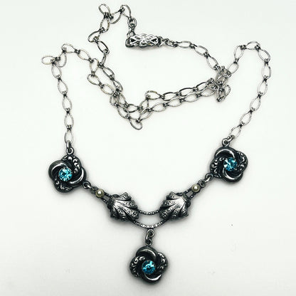 Close up of vintage silver-tone necklace showing dimensional floral ornaments, textured chain and central pendant.
