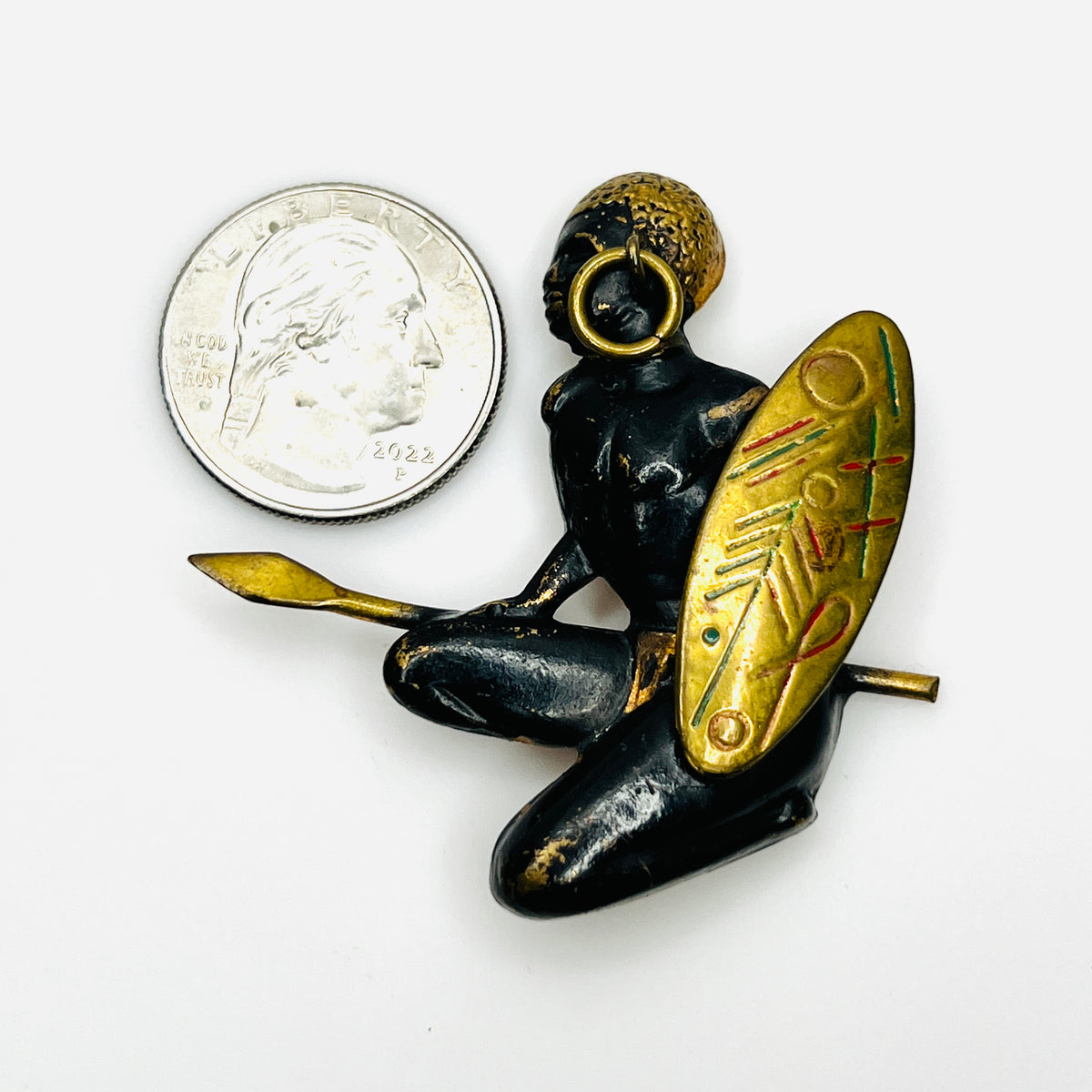 Vintage African warrior brooch beside a quarter showing its two-inch sculptural design and decorated shield.