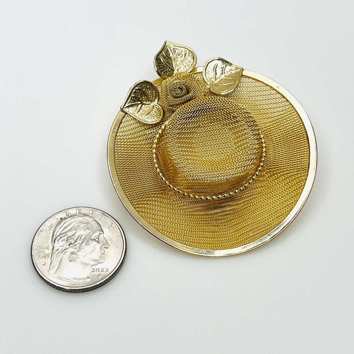 Gold mesh vintage hat brooch beside quarter showing circular brim, raised crown and floral accents