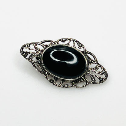 Vintage sterling silver brooch with oval black onyx cabochon, marcasite accents, and ornate filigree setting.